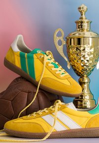 Baskets vintage jaunes et vertes, un ballon de football en cuir marron et un grand trophée en or sur un fond dégradé bleu et rose.