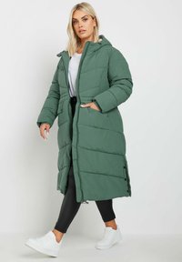 Vadderad lång kappa i mörkgrön, med huva, dragkedja och sidofickor. Kombinerad med svarta leggings och vita sneakers.