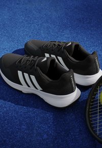 Ζευγάρι ασπρόμαυρα παπούτσια τένις Adidas Gamecourt 2 σε μπλε επιφάνεια γηπέδου δίπλα σε ρακέτα τένις με κίτρινη μπάλα.