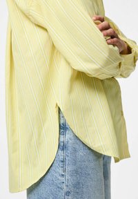 Camicia gialla a righe con orlo curvo e vestibilità ampia, realizzata in tessuto leggero. Abbinata a denim azzurro chiaro.