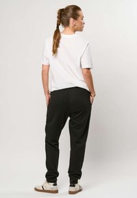 Kvinne med flettehår iført hvit kortermet skjorte, svarte bukser, store øredobber og hvite joggesko, stående med ryggen mot oss.