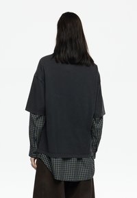 Camiseta gris carbón de gran tamaño con mangas cortas, superpuesta sobre una camisa con patrón de cuadros verde y azul marino. Corte relajado y dobladillo redondeado.