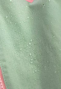 Tessuto resistente all'acqua di un verde morbido, con gocce sulla superficie. Un accento rosa è visibile sul bordo. Texture liscia con lucentezza.