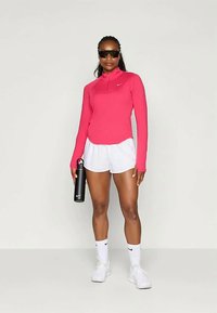 Hosszúujjú rózsaszín sportpóló, cipzáros gallérral, fehér rövidnadrággal párosítva. Fekete műanyag vízpalack és fehér sportcipő fekete részletekkel.