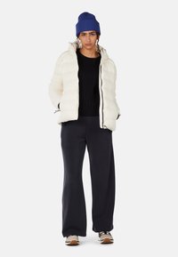 Off-white Pufferjacke mit gestepptem Design, Kapuze und schwarzem Reißverschluss; kombiniert mit einem schwarzen gerippten Pullover und lässigen, maritimen Hosen. Blauer Strickmütze getragen.