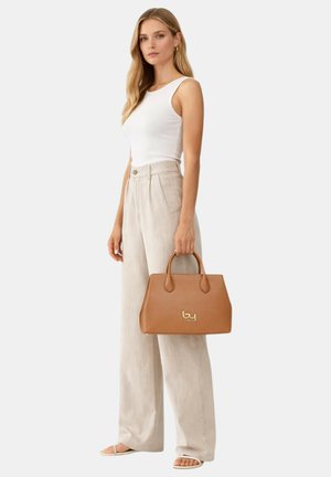 Femme portant un haut blanc sans manches et un pantalon large beige, tenant un sac à main en cuir marron avec un logo doré.