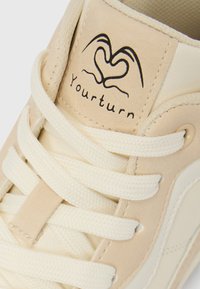 Μπεζ αθλητικό παπούτσι με λευκά κορδόνια, υφασμένο δερμάτινο επάνω μέρος και κεντημένο λογότυπο που γράφει «Yourturn» πάνω από ένα σχέδιο καρδιάς στη γλώσσα.