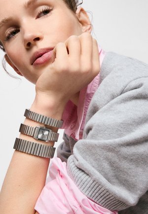 Joven mujer apoyando su barbilla en la mano, vistiendo un suéter gris, camisa rosa y tres pulseras de metal plateado, una con esfera cuadrada.
