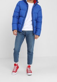 Blauwe pufferjack met capuchon, rode binnenvoering, ritssluiting. Witte t-shirt, blauwe spijkerbroek en rode hoge sneakers maken de outfit compleet.