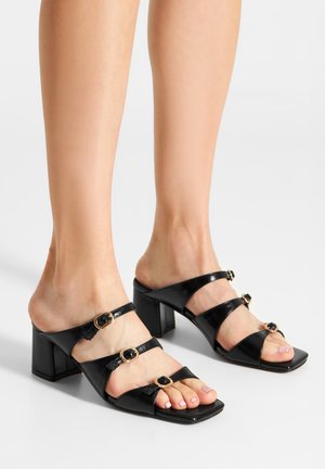 Black patent leather block-heeled sandals featuring two toe straps and two ankle straps, adorned with circular gold accents.