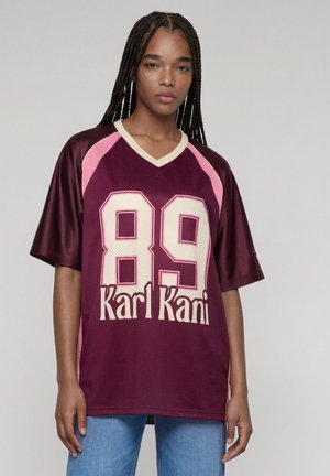 Jeune personne aux longs cheveux tressés porte un maillot de sport grenat et rose avec le numéro 89 et le texte "Karl Kani", associé à un jean bleu.