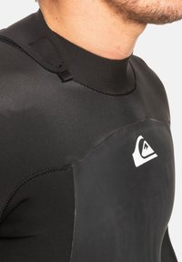 Zwart wetsuit met een hoge hals, voorzien van een textuurpaneel op de borst. Inclusief een kleine connector bij de hals en een witte logo-detail op de borst.