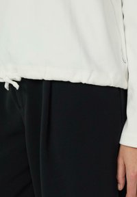 Witte fleece top met een trekkoord aan de zoom, gecombineerd met zwarte onderkleding; gladde textuur en casual ontwerp.