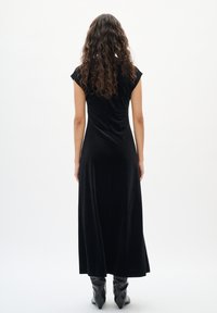 Frau mit langen lockigen Haaren, die ein schwarzes, kurzes Maxikleid und schwarze spitze Stiefel trägt, steht mit dem Rücken zu einer weißen Wand.