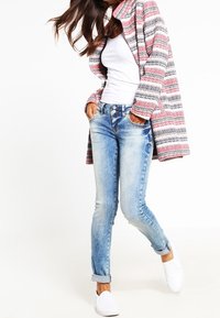 Kvinna i vit linne, randig rosa och grå kardigan, ljusblå jeans med uppvikta ben och vita slip-on skor, som står med händerna i fickorna.