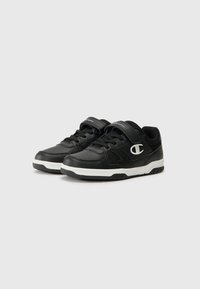 Chaussures de sport noires avec une tige en synthétique, dotées d'un système de fermeture à lacets et d'une sangle Velcro, d'accents de semelle blanche et de détails texturés.