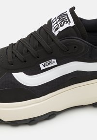 Baskets Vans noires avec une rayure latérale blanche, lacets noirs et semelle beige, affichant des étiquettes de logo sur la languette et le panneau latéral.