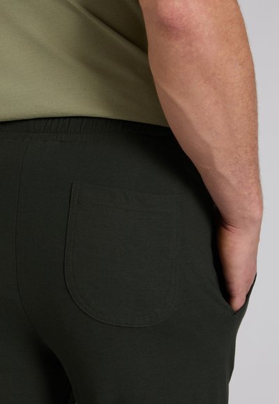 Mężczyzna w czarnych spodniach z tylną kieszenią z naszywką i boczną kieszenią na biodrze, ręka spoczywająca w bocznej kieszeni, widoczne przedramię i zielony brzeg koszuli.