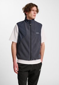 Junger Mann in einer dunkelgrauen Reißverschluss-Fleeceweste über einem weißen kurzärmeligen Hemd und schwarzen Hosen, der mit den Händen in den Taschen steht und nach rechts schaut.