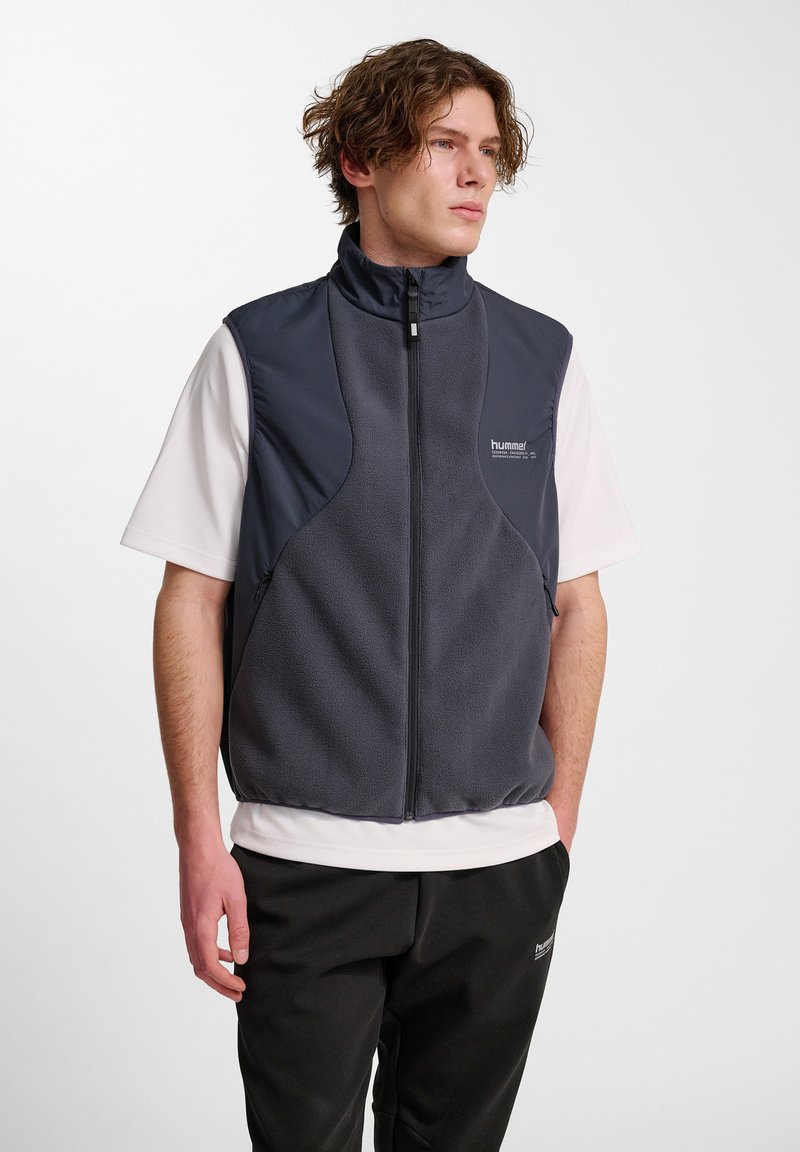 Junger Mann in einer dunkelgrauen Reißverschluss-Fleeceweste über einem weißen kurzärmeligen Hemd und schwarzen Hosen, der mit den Händen in den Taschen steht und nach rechts schaut.