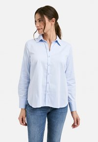 Woman wearing a light blue button-up shirt with long sleeves and blue jeans, looking to her left against a plain white background.