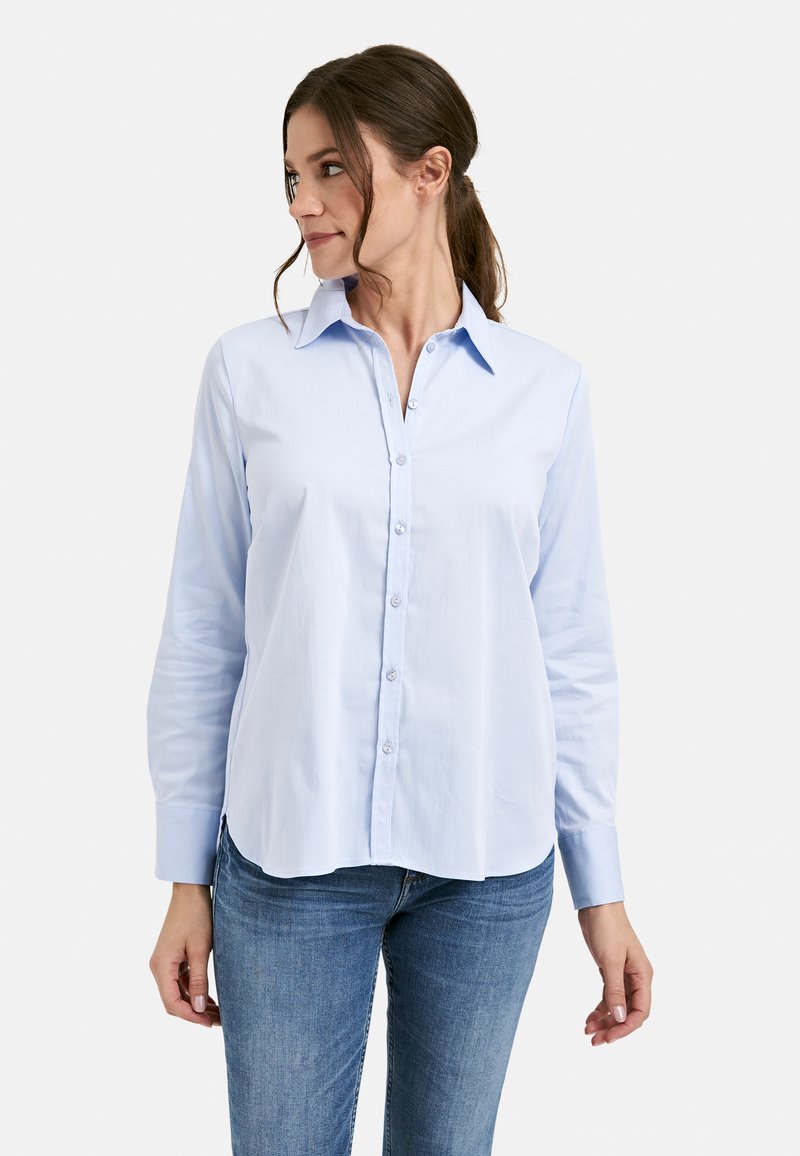 Woman wearing a light blue button-up shirt with long sleeves and blue jeans, looking to her left against a plain white background.