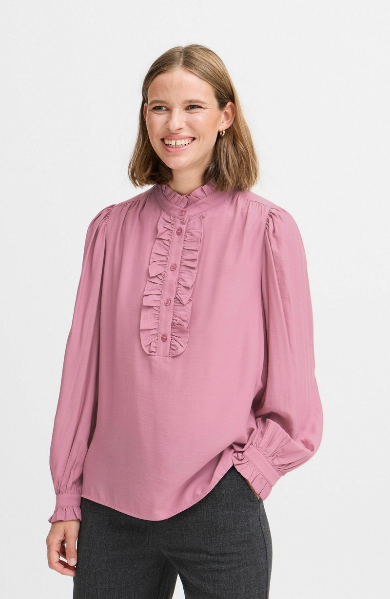 Blusa rosa con un alto cuello rizado, mangas largas abullonadas y un frente abotonado que presenta un panel rizado en el centro. Tejido suave y liso.