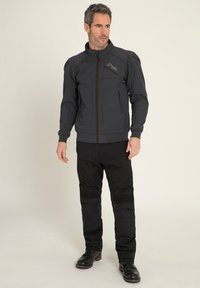 Giacca zip nera scura con colletto alto, dettagli sulle tasche e accenti rossi. Abbinata a pantaloni neri con sezioni delle ginocchia a coste.