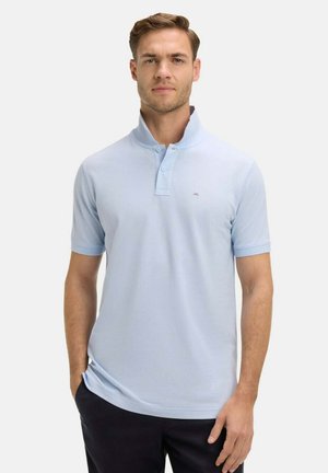 Homme portant un polo bleu clair à manches courtes avec le col relevé, debout devant un fond uni, une main dans la poche.