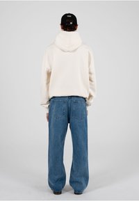 Persoană purtând un hanorac crem, blugi albaștri largi și o șapcă neagră, văzută din spate, pe un fundal simplu gri deschis.