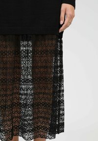 Jupe en dentelle noire avec un motif en zigzag et des détails floraux. La dentelle recouvre une base en tissu solide, créant un effet de texture en couches.