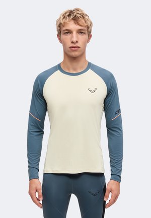Junger Mann mit kurzem blondem Haar, der ein cremefarbenes und blaues langärmeliges Sportshirt sowie passende blaue Leggings trägt und nach vorne schaut.