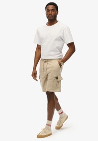 Pantaloni cargo beige con tasche laterali, vita elastica e dettaglio con cordino, abbinati a una maglietta bianca e sneakers beige.