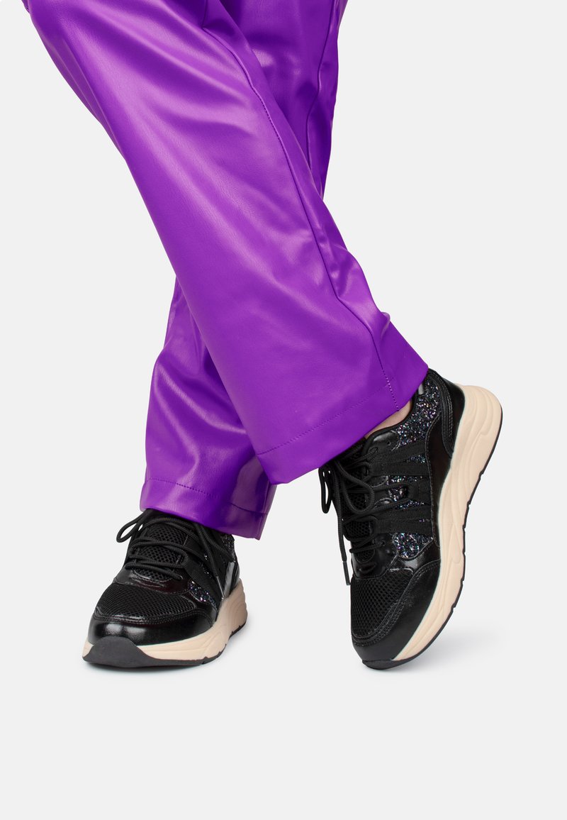 Zwarte sneakers met een gestructureerd oppervlak en reflecterende accenten, gecombineerd met glanzende paarse broek met een gladde afwerking.