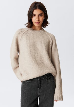 Femme aux cheveux bruns portant un pull texturé beige et un jean noir taille haute, se tenant devant un fond clair.