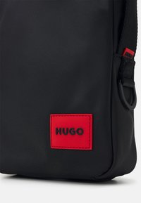 Borsa in nylon nera con un evidente patch logo in gomma rossa con la scritta "HUGO." Texture liscia con un design elegante e hardware laterale robusto.