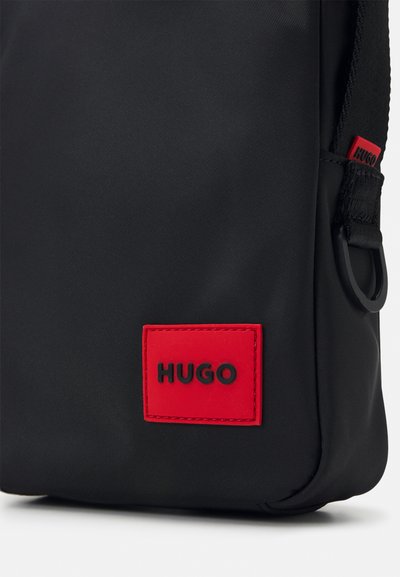 Sac en nylon noir avec un patch en caoutchouc rouge bien en vue portant l'inscription "HUGO." Texture lisse, design épuré et matériel latéral robuste.
