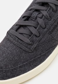 Närbild av en mörkgrå strukturerad sneaker med matchande snörning och vit sula, som framhäver sömmar och tygdetaljer.