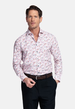 Man met kort bruin haar, gekleed in een wit bloemenshirt met knopen, donkere broek en een bruine riem, poseert met één hand in de zak.