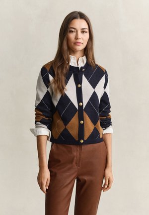 Femme portant un cardigan boutonné à motif losanges bleu, blanc et marron sur une chemise blanche avec un pantalon marron, debout devant un fond neutre.