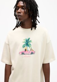 Beżowa oversize koszulka t-shirt z graficznym nadrukiem różowego kabrioletu, palm i napisem "Miami" w różowym, odręcznym kroju pisma. Krótkie rękawy, miękka tekstura.