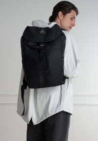 Zaino nero con un cappuccio arrotondato, cinturino anteriore con fibbia e etichetta del marchio. Realizzato in materiale resistente, presenta spallacci regolabili e tasche laterali.