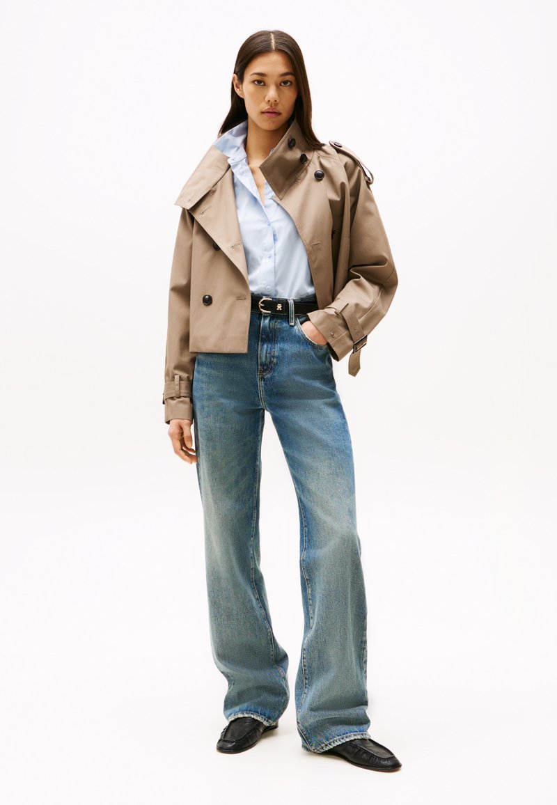 Beige trenchcoat met grote zwarte knopen, gedragen over een lichtblauw overhemd en een hoge taille blauwe jeans, afgewerkt met zwarte schoenen.