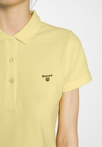 Polo shirt jaune en tissu texturé avec une patte de boutonnage à trois boutons, présentant un logo noir brodé sur la zone de la poitrine gauche.