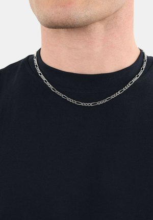 Nahaufnahme einer Person, die eine silberne Ketten-Halskette über einem schwarzen Rundhals-Shirt gegen einen schlichten Hintergrund trägt.