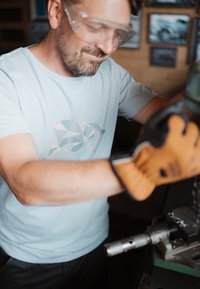 Camiseta de algodón azul claro con un diseño geométrico de un oso polar, usada con guantes acolchados marrones, mientras maneja un taladro de maquinaria.