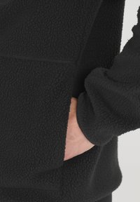 Pull en polaire noir avec un extérieur texturé, comportant une poche frontale et des poignets côtelés, mettant en avant la chaleur et le confort.