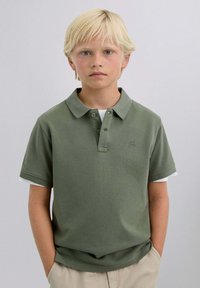 Polo oliva con un tejido texturizado, botonera de dos botones y logo bordado en el pecho. Llevado sobre una camiseta blanca.