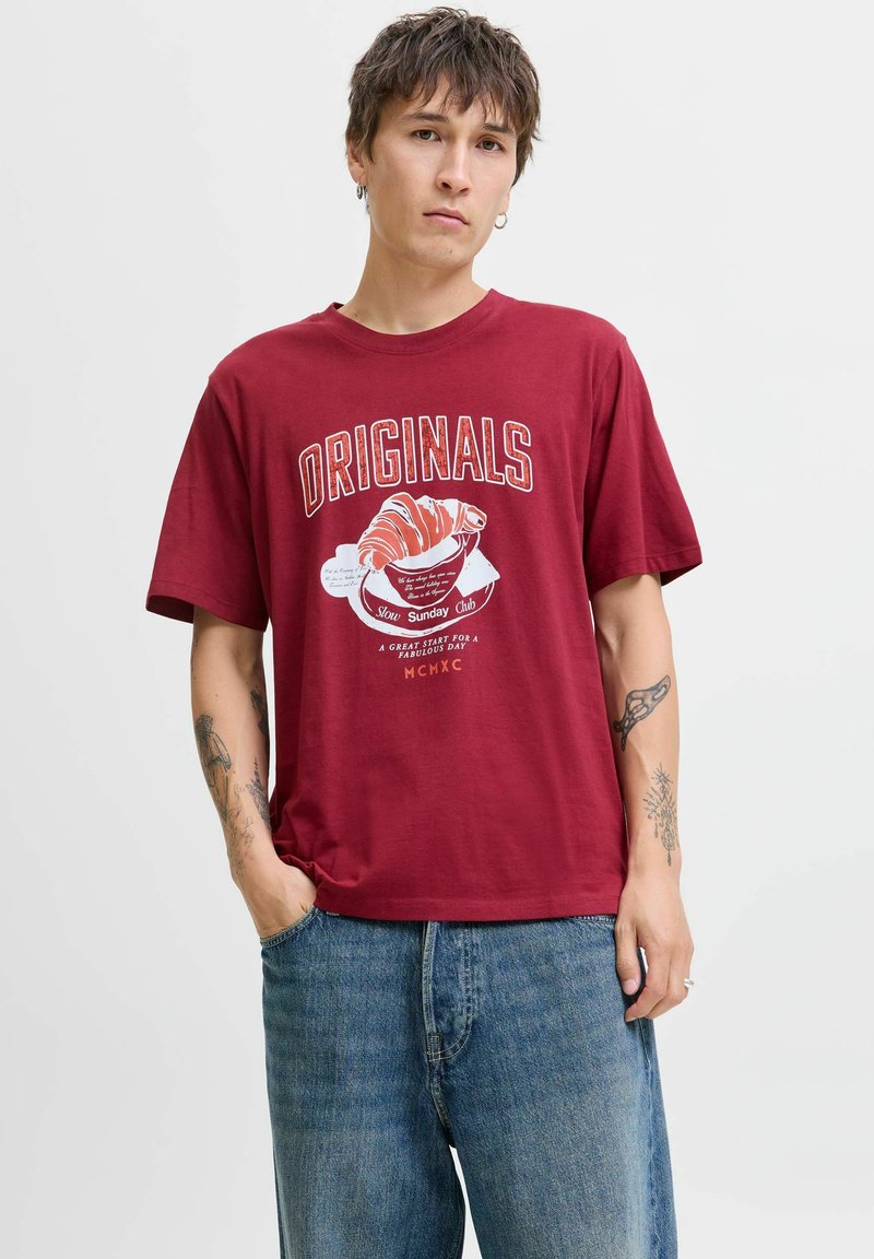 Rode katoenen t-shirt met een grafische print van een croissant en de tekst "ORIGINALS." Relaxte pasvorm, korte mouwen, gedragen met blauwe denim jeans.