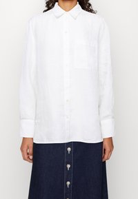 Biała lniana koszula zapinana na guziki z klasycznym kołnierzem, długimi rękawami i jedną kieszenią na piersi. Noszona z dżinsową spódnicą z guzikami.